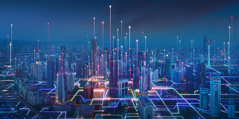 "A digital cityscape visualization showing a nighttime urban skyline with skyscrapers and high-rise buildings illuminated by glowing blue and purple lights. The image is overlaid with a network of colorful digital connection lines, data points, and vertical light beams extending upward from various buildings, symbolizing IoT connectivity, smart city infrastructure, and data transmission in an interconnected metropolitan environment."